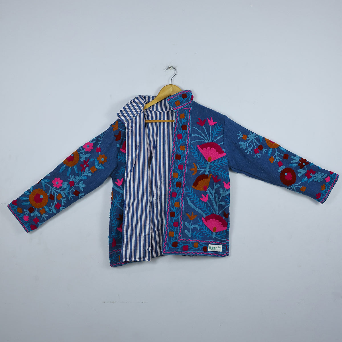 A blue embroidered jacket with colorful floral patterns and a striped inner lining, hanging on a wooden hanger against a plain light gray wall.